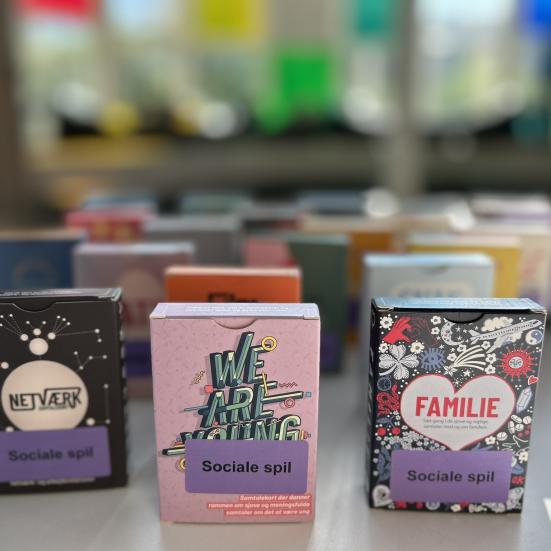 Samtalespil på størrelse med en pakke spillekort med forskellige titler står på et bord. Baggrunden viser slørede farver.