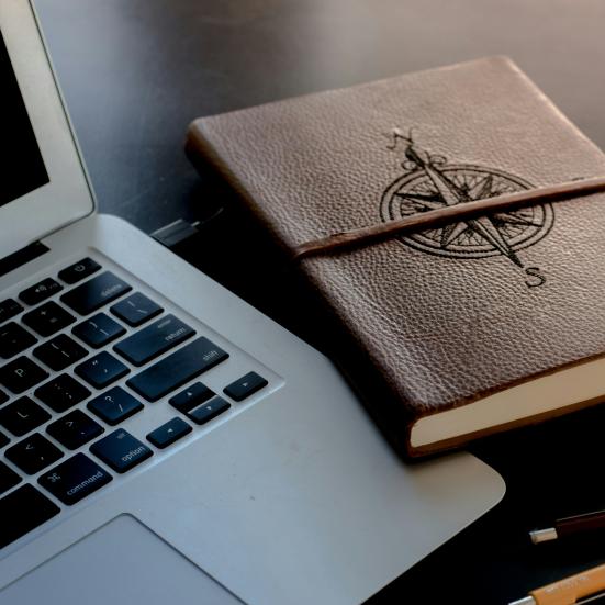 Billede af en brun notesbog i læder med et kompas påtrykt på forsiden. I venstre side af billedet anes en computer og i højre nederste side ses 2 træblyanter