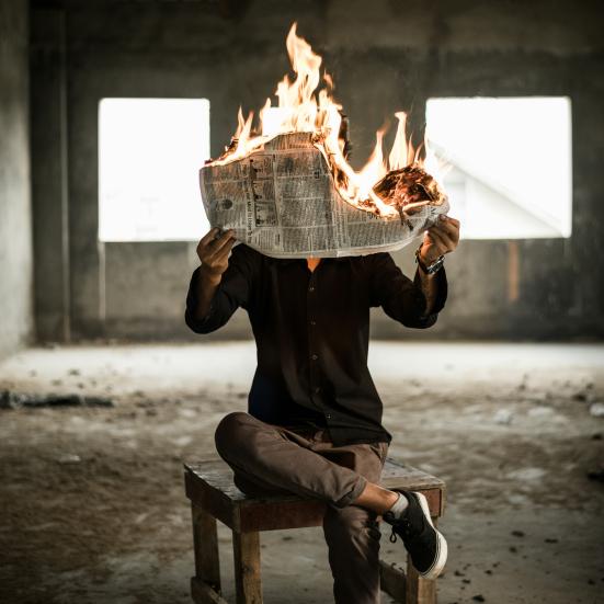 Person sidder på en stol med benene over kors og holder en brændende avis op for an sig.