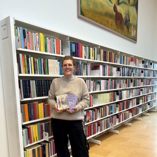 Michelle står på biblioteket. Hun holder "Øvelser i mørke" og "Poesibog" i hænderne og kigger smilende ind i kameraet. Bag Michelle ses fyldte bogreoler og et stort maleri på væggen.
