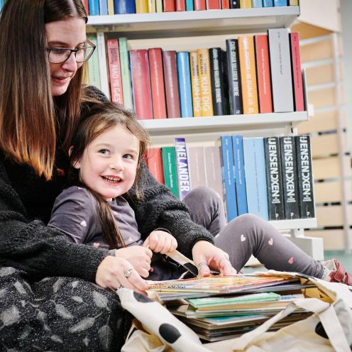 En voksen og et barn sidder i et bibliotek og kigger i en bog sammen
