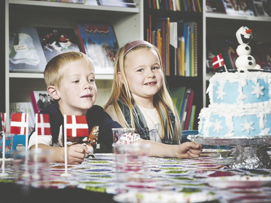 To børn sidder ved et bord og smiler. På bordet står der fødselsdagsflag og en kage.