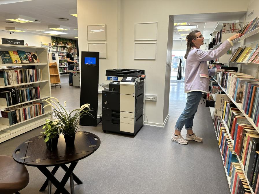 Printeren på Svenstrup Bibliotek står i midten af billedet. I højre side står en kvinde og rækker ud efter en bog på hylden.