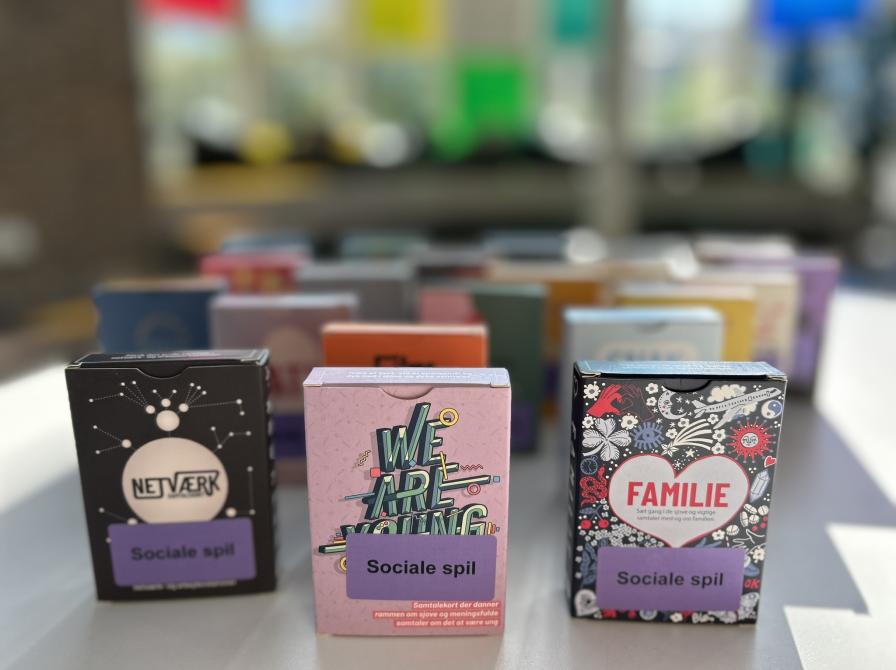 Samtalespil på størrelse med en pakke spillekort med forskellige titler står på et bord. Baggrunden viser slørede farver.