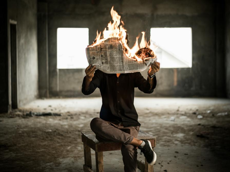 Person sidder på en stol med benene over kors og holder en brændende avis op for an sig.