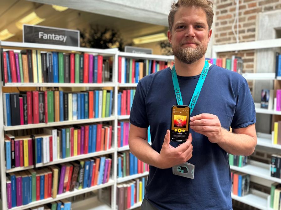 Allan står foran fantasy-reolen på Hovedbiblioteket. Han holder sin telefon foran sig, hvor han viser lydbogsudgaven af hans anbefaling.