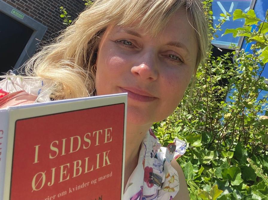 Marianne sidder udendørs og læser i bogen "I sidste øjeblik" foran Glashjørnet på Hovedbiblioteket.