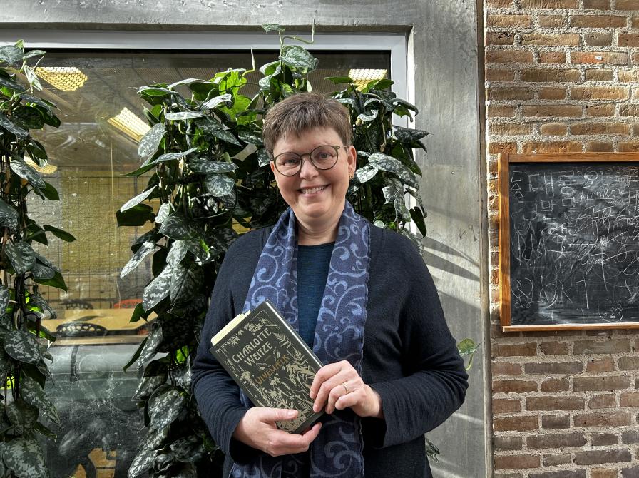 Vita står foran de grønne planter foran Makerspace på Hovedbiblioteket. Hun smiler og holder bogen "Ulvemælk" i hænderne. Lyset falder flot på væggen bag hende.