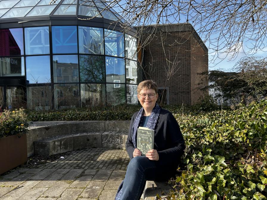 Vita sidder i bibliotekshaven ved Hovedbiblioteket. Bag hendes ses glashjørnet med dets farverige ruder. Vita smiler og himlen er blå.